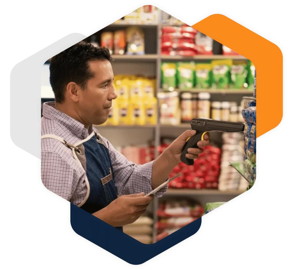 A man in a checkered shirt and apron uses a handheld barcode scanner to scan products on a shelf in a grocery store. Colorful packaged items are visible in the background. Contact us for more information.