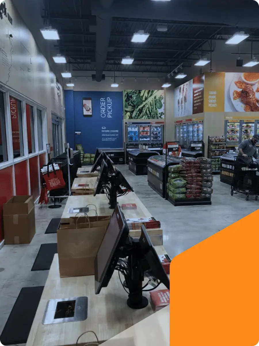 A grocery store checkout area with unattended registers, paper bags on the counter, self-checkout screens, and shelves of produce and packaged goods. Signs indicate order pickup and bakery sections.
