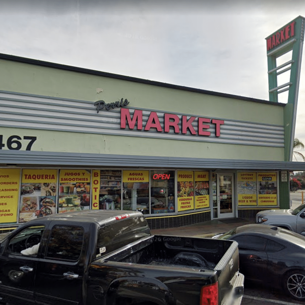 A storefront with a large sign reading Perus MARKET. Bright yellow posters in the windows advertise food and beverages. Several vehicles are parked in front of the store, and a tall sign also reads MARKET.
