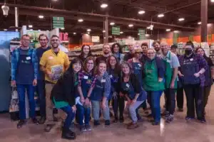 A group of grocery store employees in aprons and name tags pose together inside a supermarket aisle, smiling at the camera under bright lights. Shelves stocked with products are visible in the background.