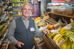 An older man with glasses and gray hair, wearing a vest and name tag, smiles while standing by a fruit display with bananas and mangoes in a grocery store aisle, where he efficiently oversees the produce using a modern grocery store inventory management system.